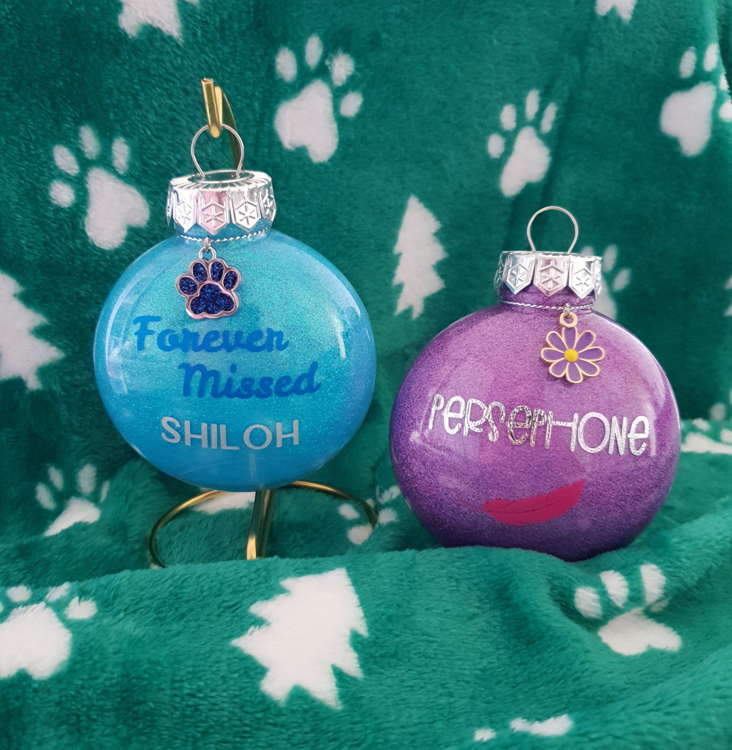 Two Christmas ornaments, one blue and one purple, with vinyl names on a green fabric background with paw prints.