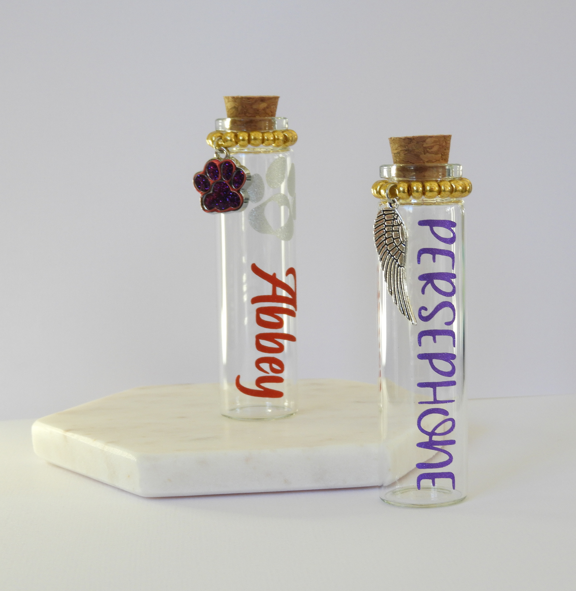 The photo displays two glass keepsake bottles with cork stoppers, charms and vinyl lettering for the pet’s name. 