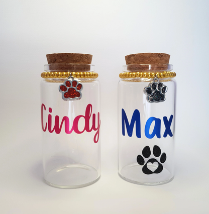 The photo displays two glass keepsake bottles with cork stoppers, charms and vinyl lettering for the pet’s name. The bottles measure 9.5x4.7cm (3.8x1.9in).