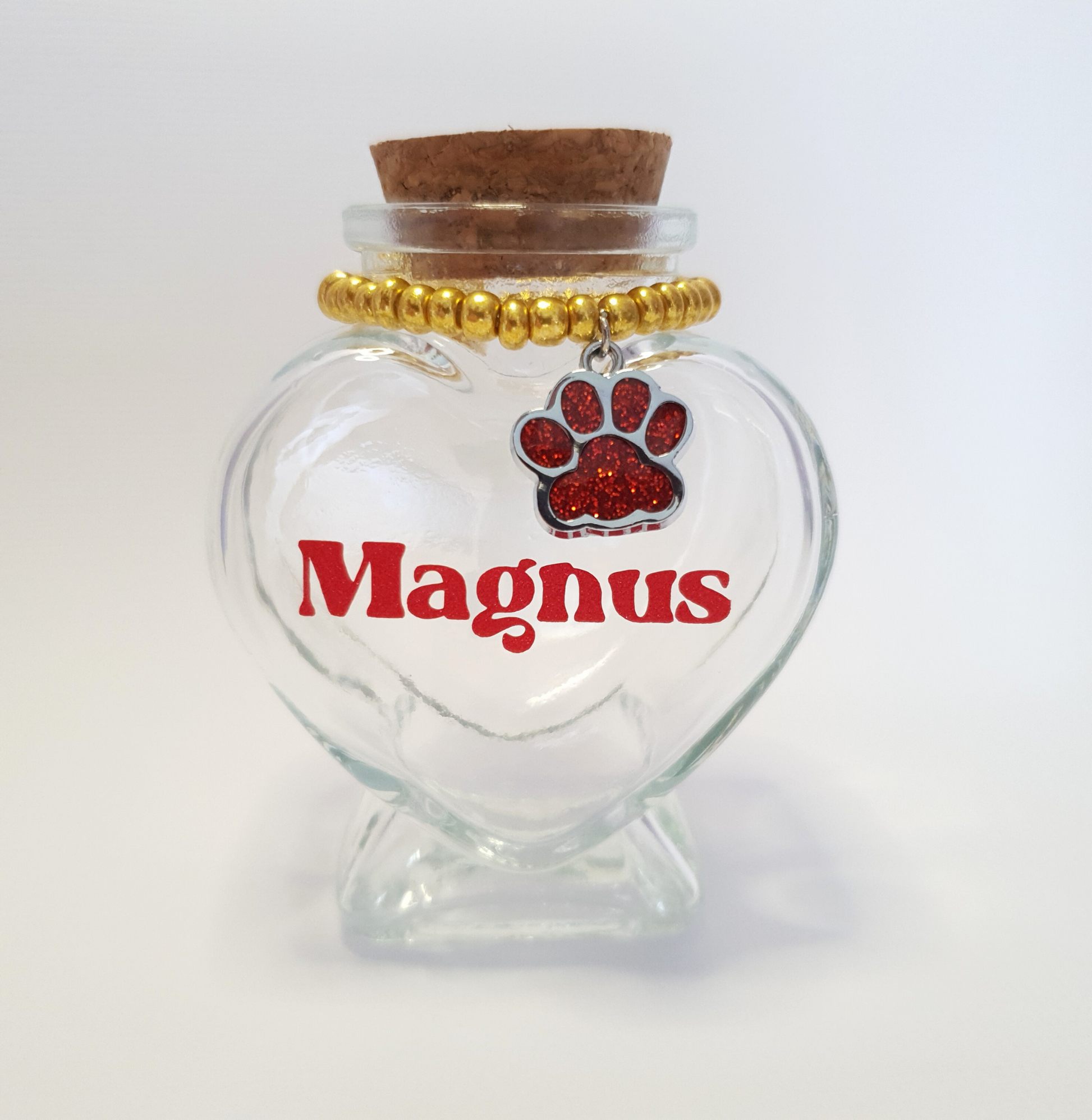 The photo displays a heart-shaped glass keepsake bottle with a cork stopper, a charm and vinyl lettering for the pet’s name.
