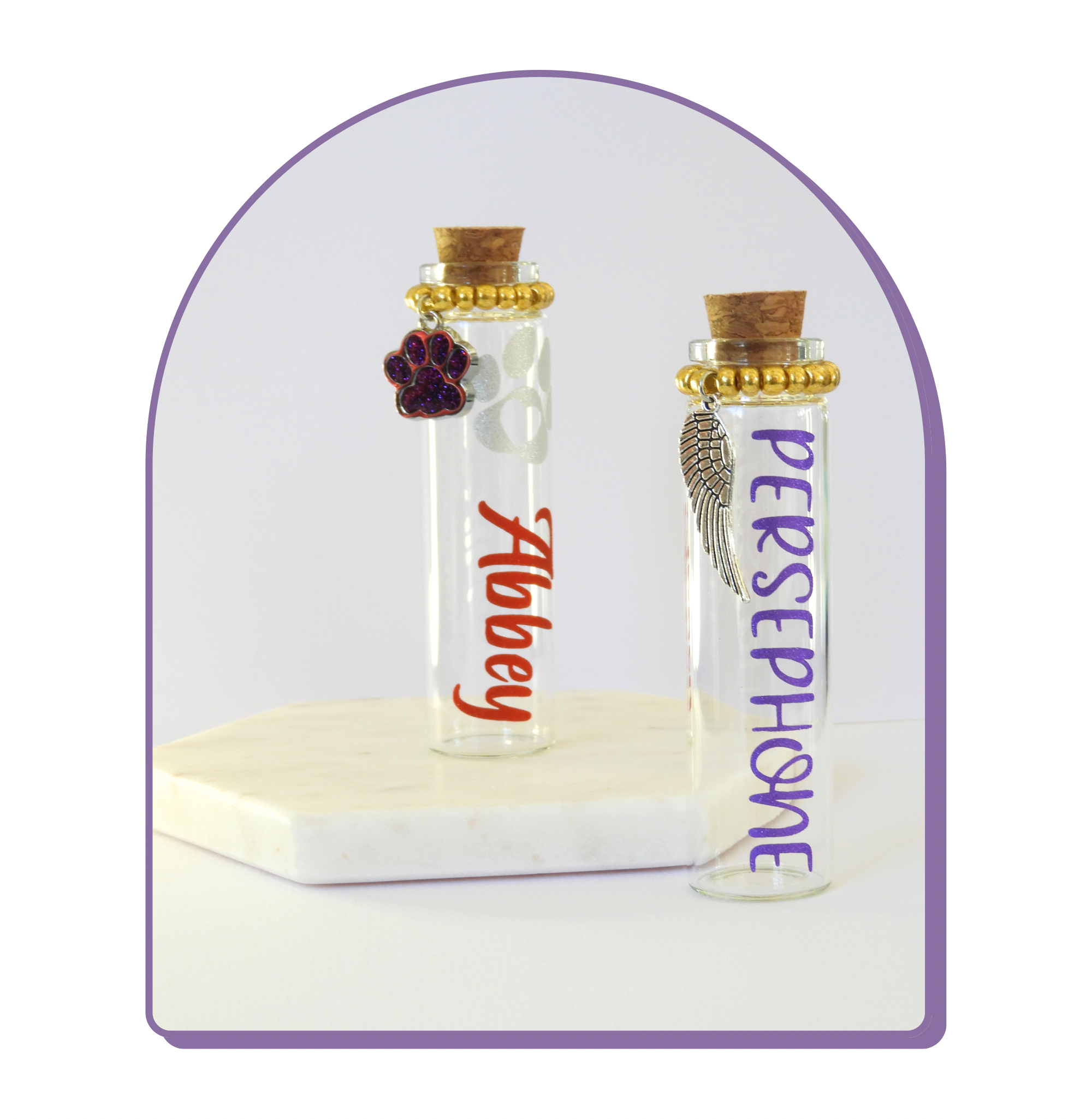 The photo displays two glass keepsake bottles with cork stoppers, charms and vinyl lettering for the pet’s name. 
