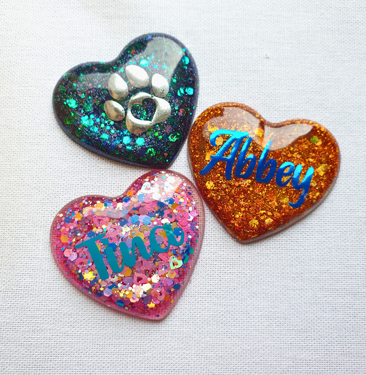 Three glittery heart-shaped resin keepsakes with names and paw prints on a white background