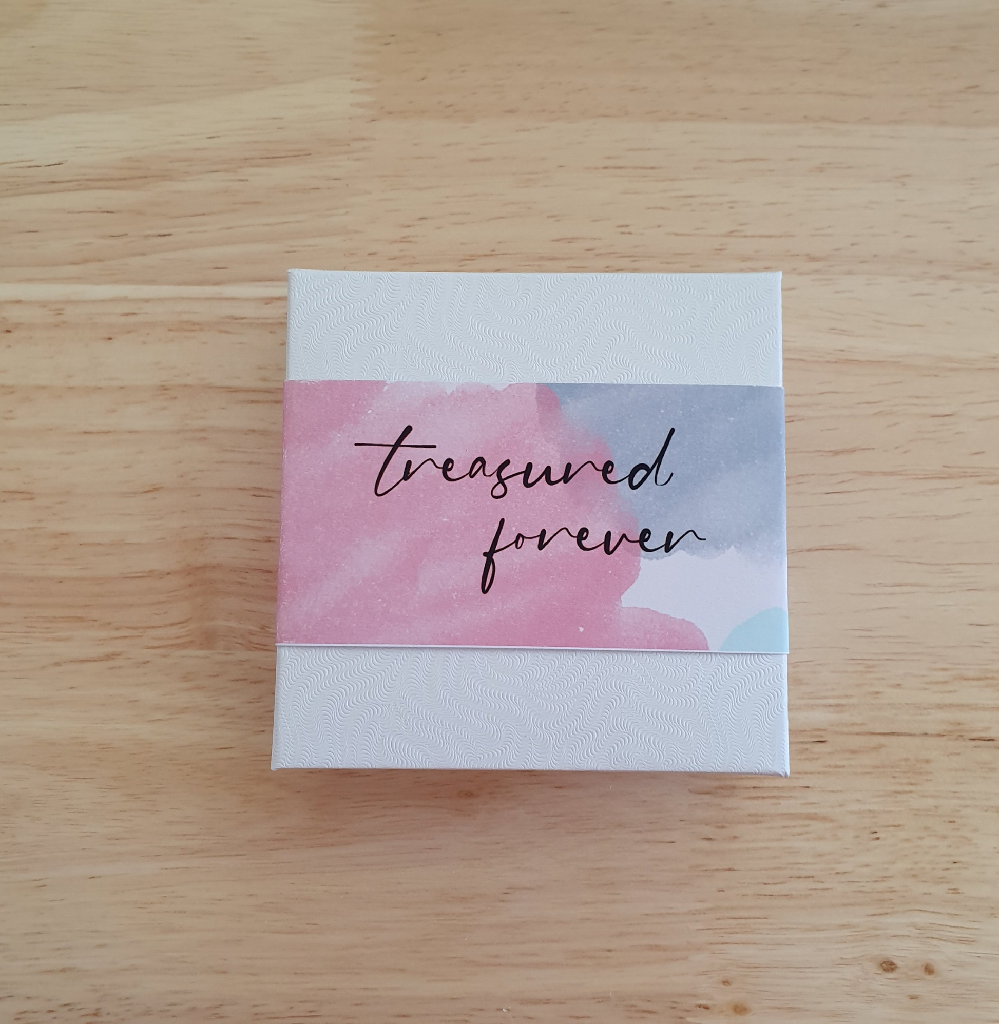 Small box with 'treasured forever' text on a watercolor background, placed on a wooden surface.